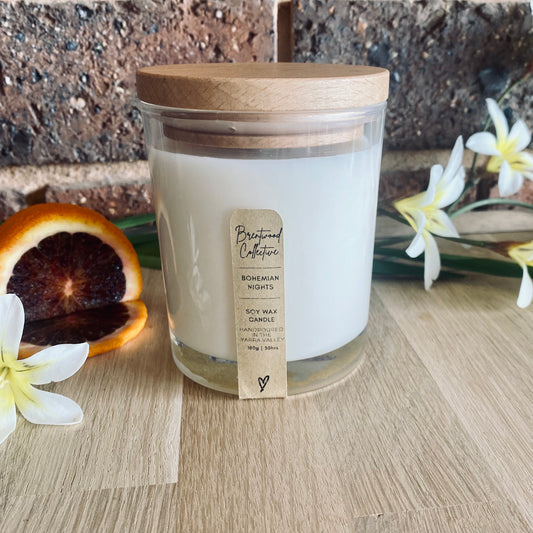 Bohemian Nights Scented Candle - clear glass jar with natural timber lid & slim/minimal Kraft brown label and black text. Set among white & yellow wildflowers and a sliced blood orange. Sitting on a natural timber bench top with brick wall in background.